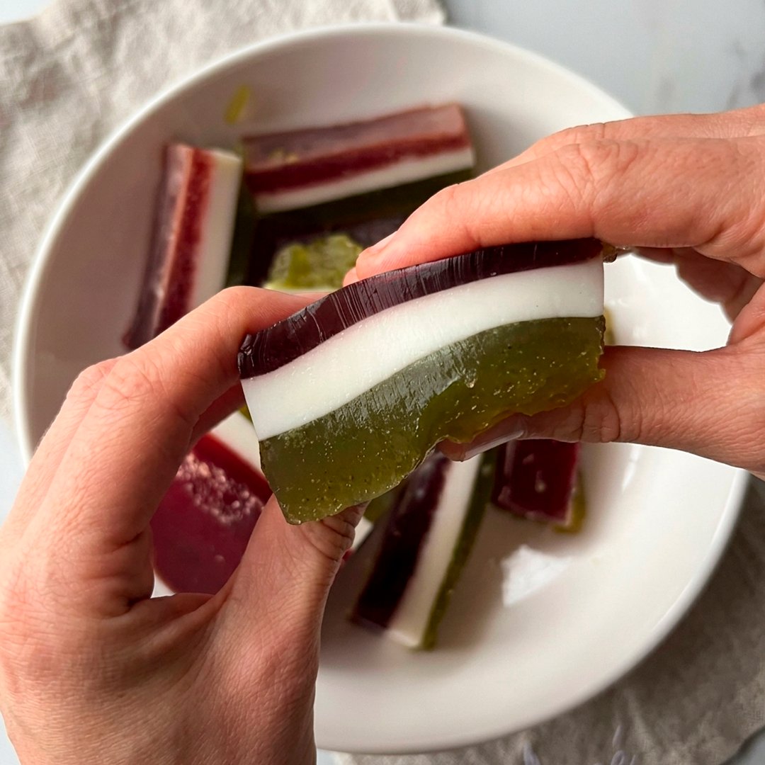 Hands holding a three-layer jelly bar showing distinct burgundy, white coconut and olive green layers, white plate with more bars below